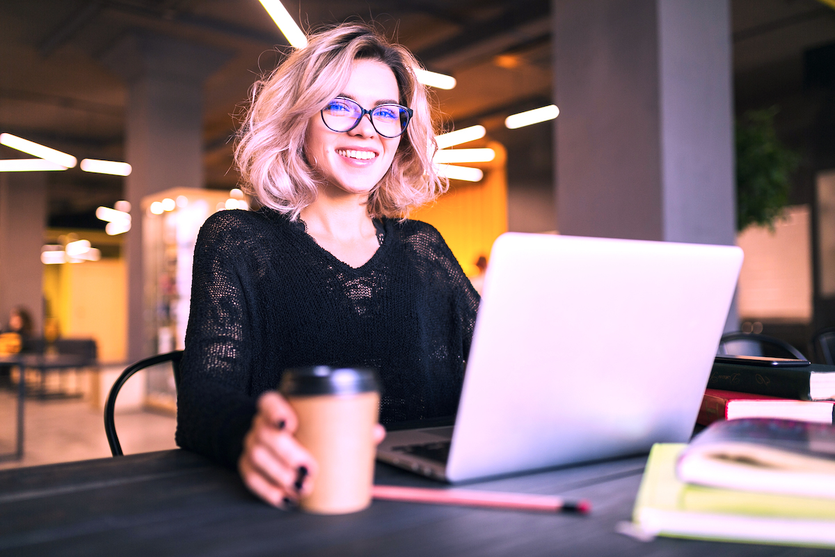 portrait of young pretty woman sitting at table in black shirt working on laptop in co-working office, wearing glasses, smiling, happy, positive, drinking coffee in paper cup